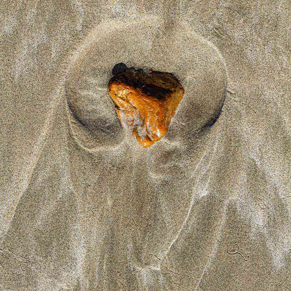 strands | A rust coloured stone cupped in sand, and shadows on a Breton beach