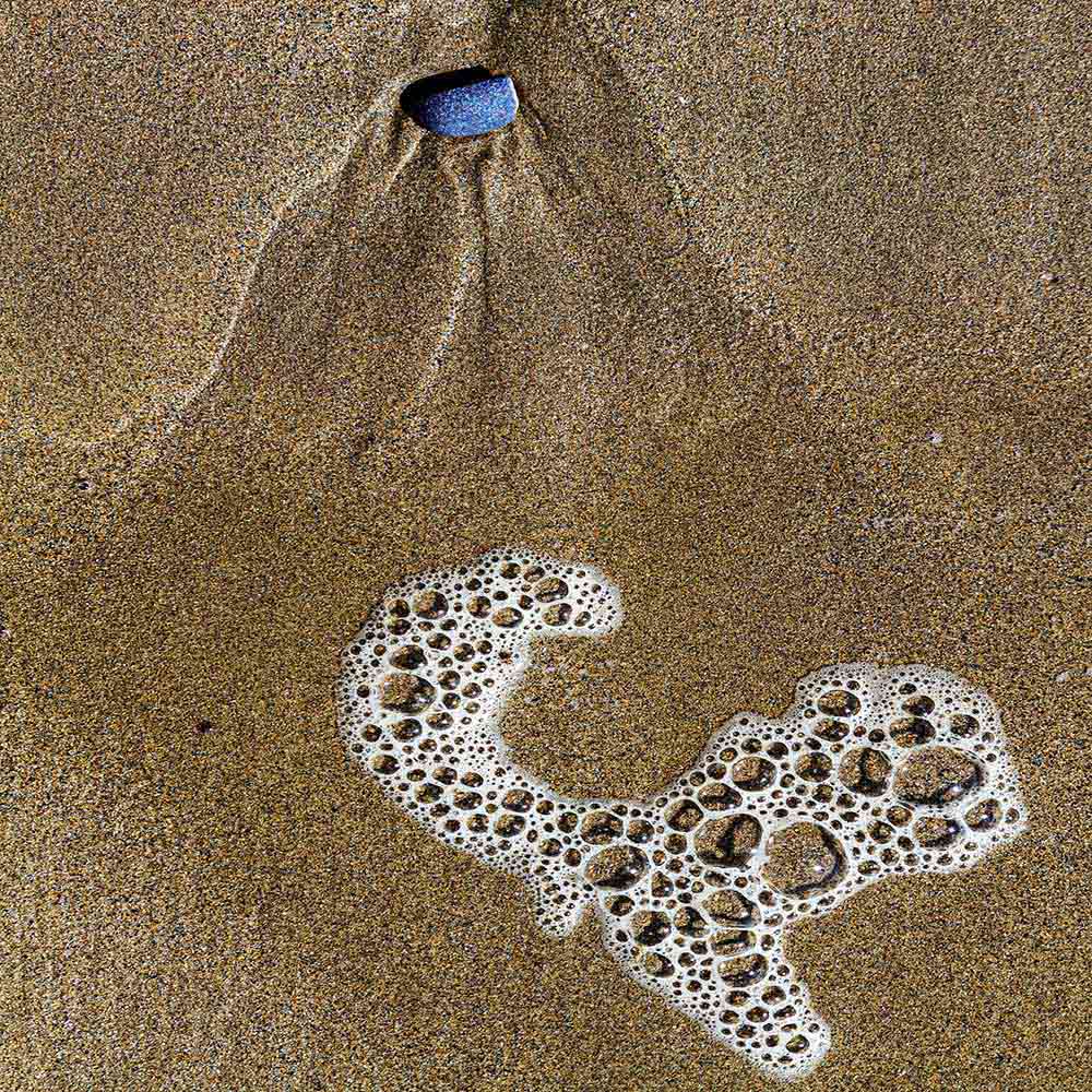 strands | A blue stone, with patterns in sand fanning out to froth, and shadows on a Breton beach