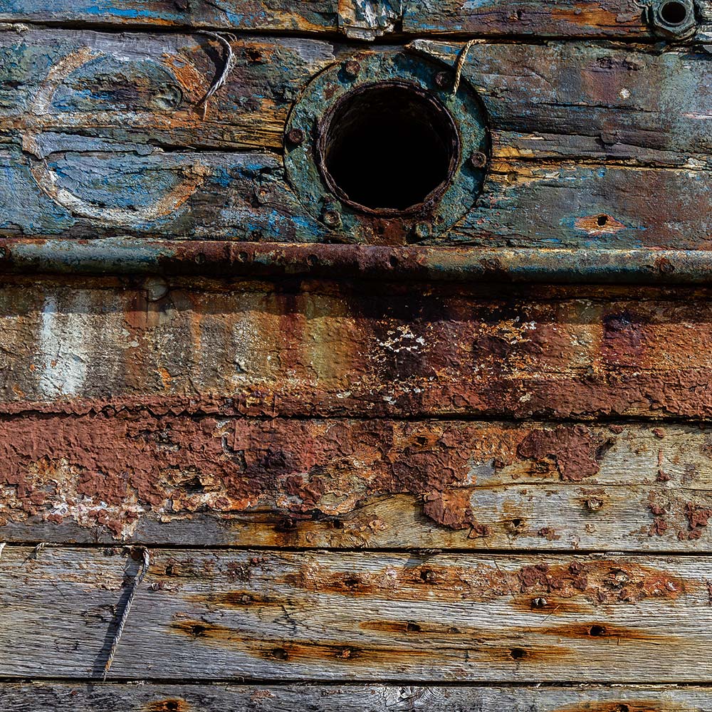 Camaret-sur_mer | The side of a burnt out rotting hulk of a fishing boat with peeling rusty white, blue and yellow paint with the design of a plimsoll line. Deeply textured worn panks of wood with loose string caulking